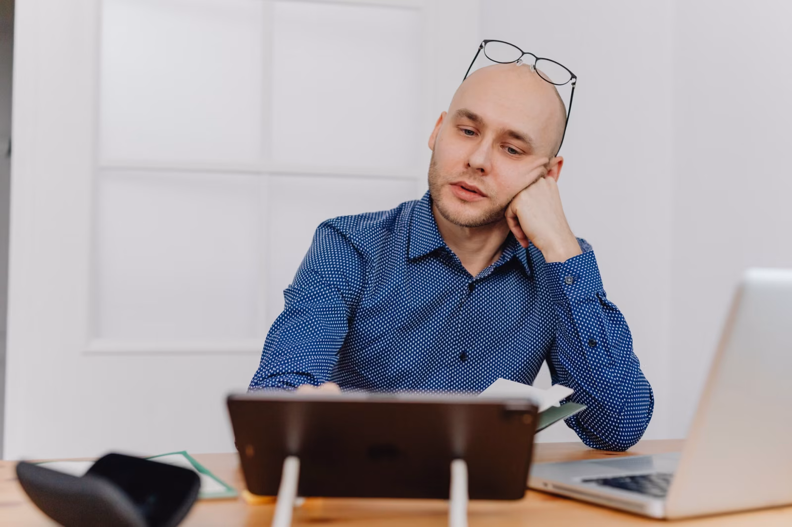 Bald man in blue shirt working at desk with laptop and tablet indoors.