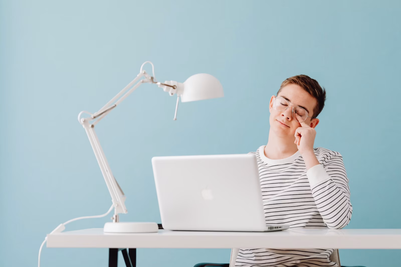 Young man looks exhausted while working on laptop at a desk with a lamp.